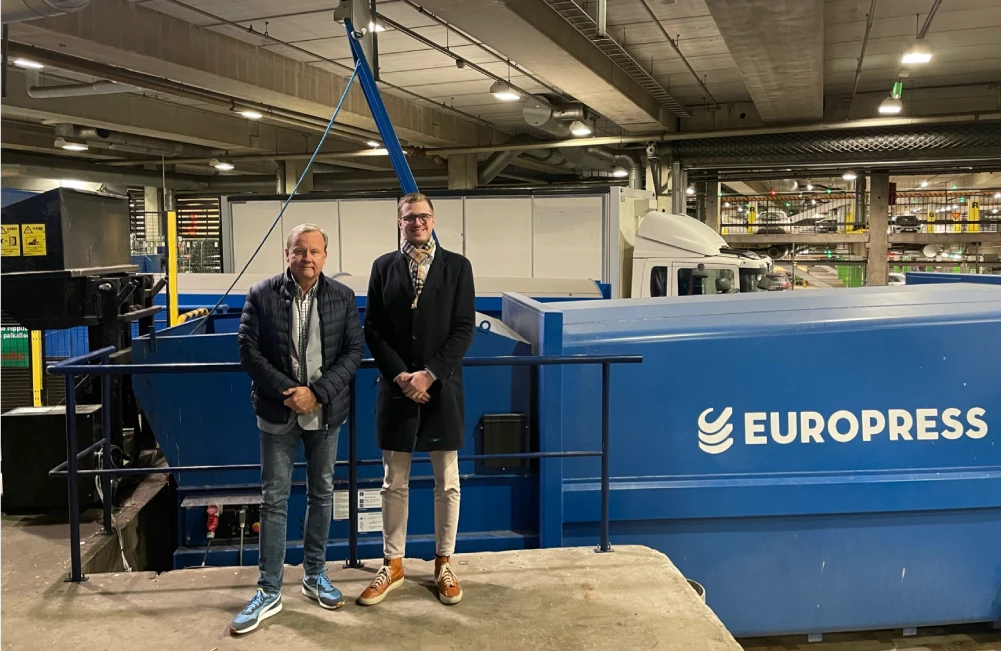 Zwei Europress Vertreter stehen vor einem großen blauen Europress Abfallverdichter in einer industriellen Recyclinganlage